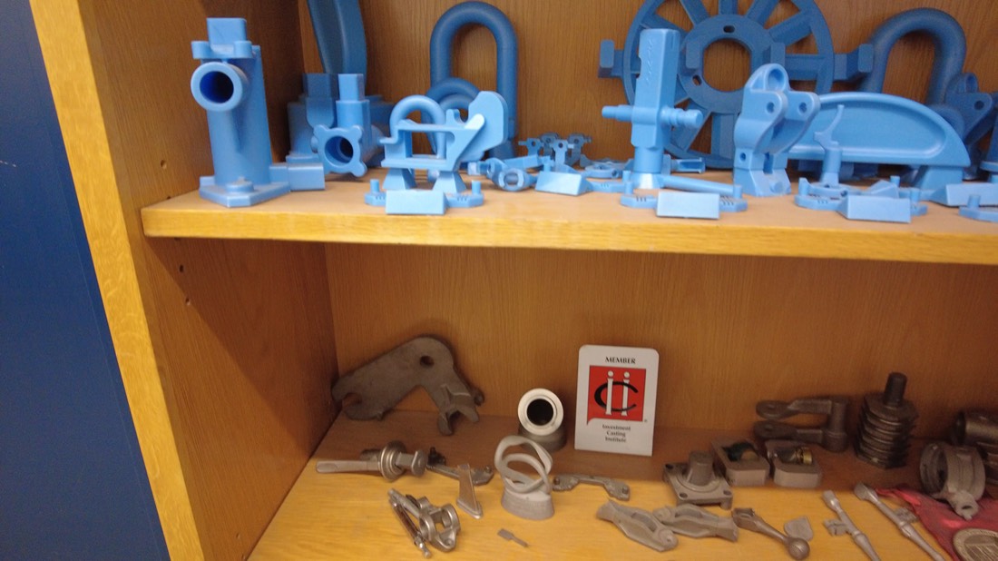 Shelf of MPI investment-cast parts: wax patterns and finished metal castings beside an Investment Casting Institute member card